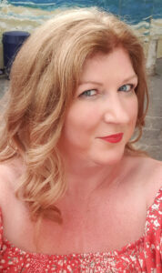 A photograph of a woman in a red dress off the shoulder. She is turning slightly away from the camera, but looking directly at it. She is smiling. She has blue eyes and red lipstick. Her hair is honey blonde, shoulder length, and curly